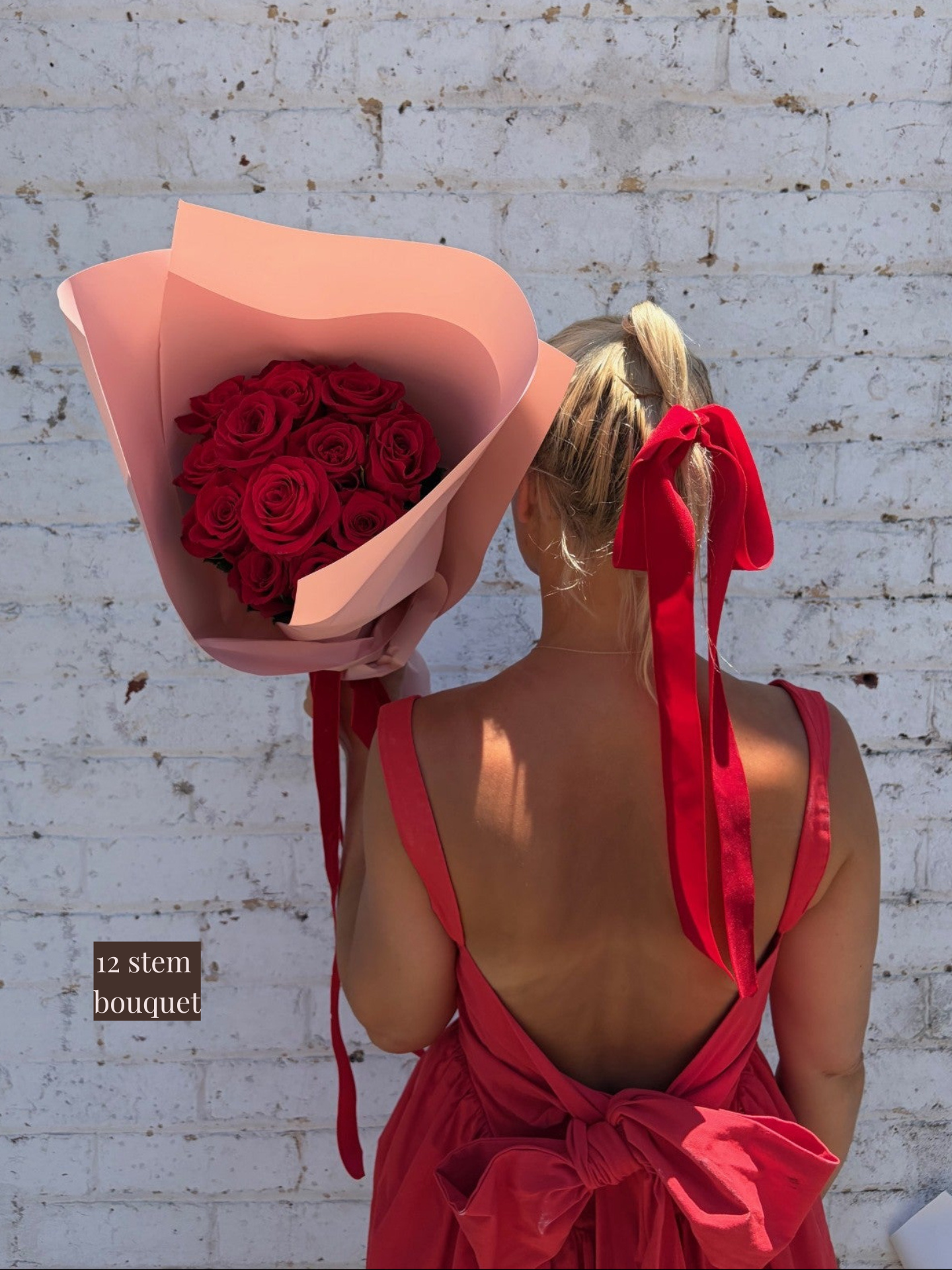 Woman in a red dress holding a large bouquet of red roses against a white brick wall.