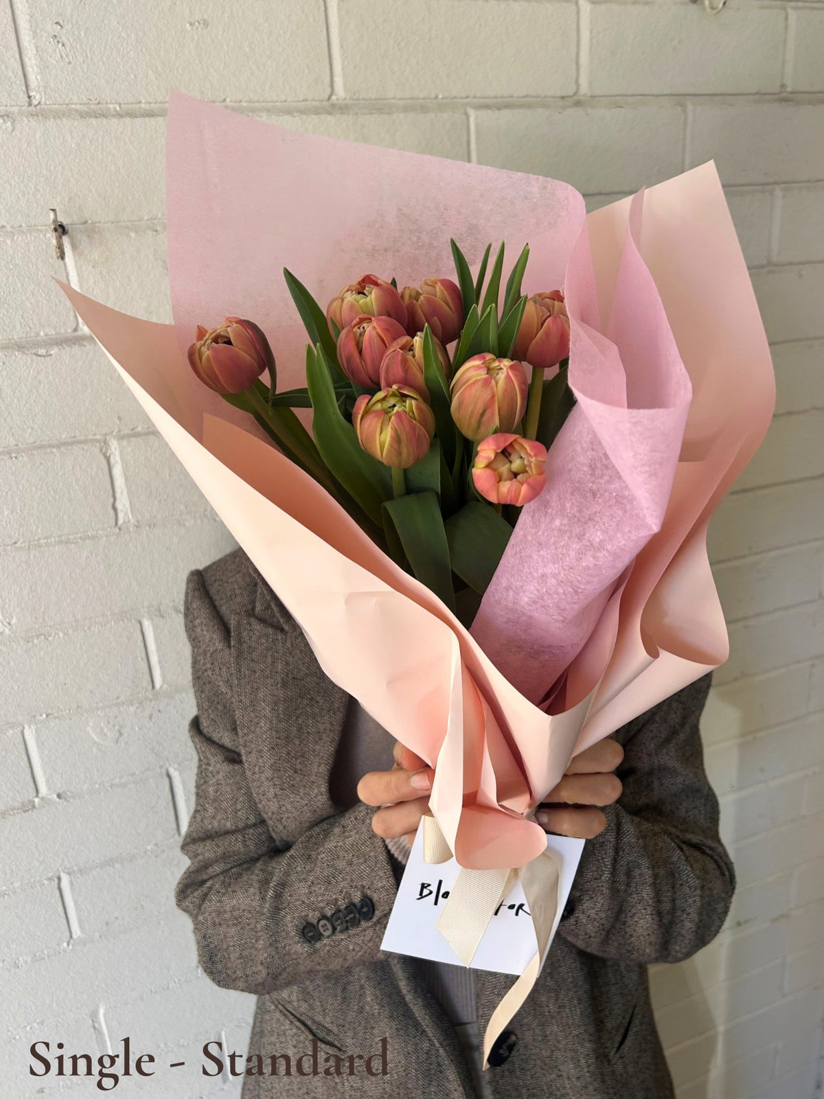 A bouquet of orange double tulips wrapped in pale pink tissue paper and a peach ribbon, held by a person wearing a gray blazer against a white painted brick wall.