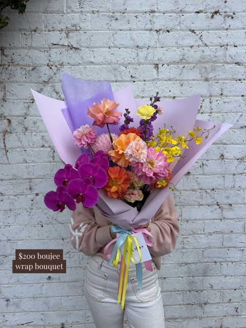 Person holding a colorful bouquet wrapped in purple paper against a white brick wall.