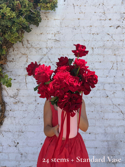 A person in a red dress holds a large bouquet of deep red roses in a striped white and red vase in front of a white brick wall. Text at the bottom reads '24 stems + Standard Vase'.