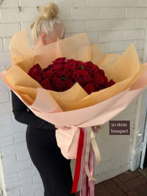 Person holding a large bouquet of red roses wrapped in brown paper against a white brick wall.