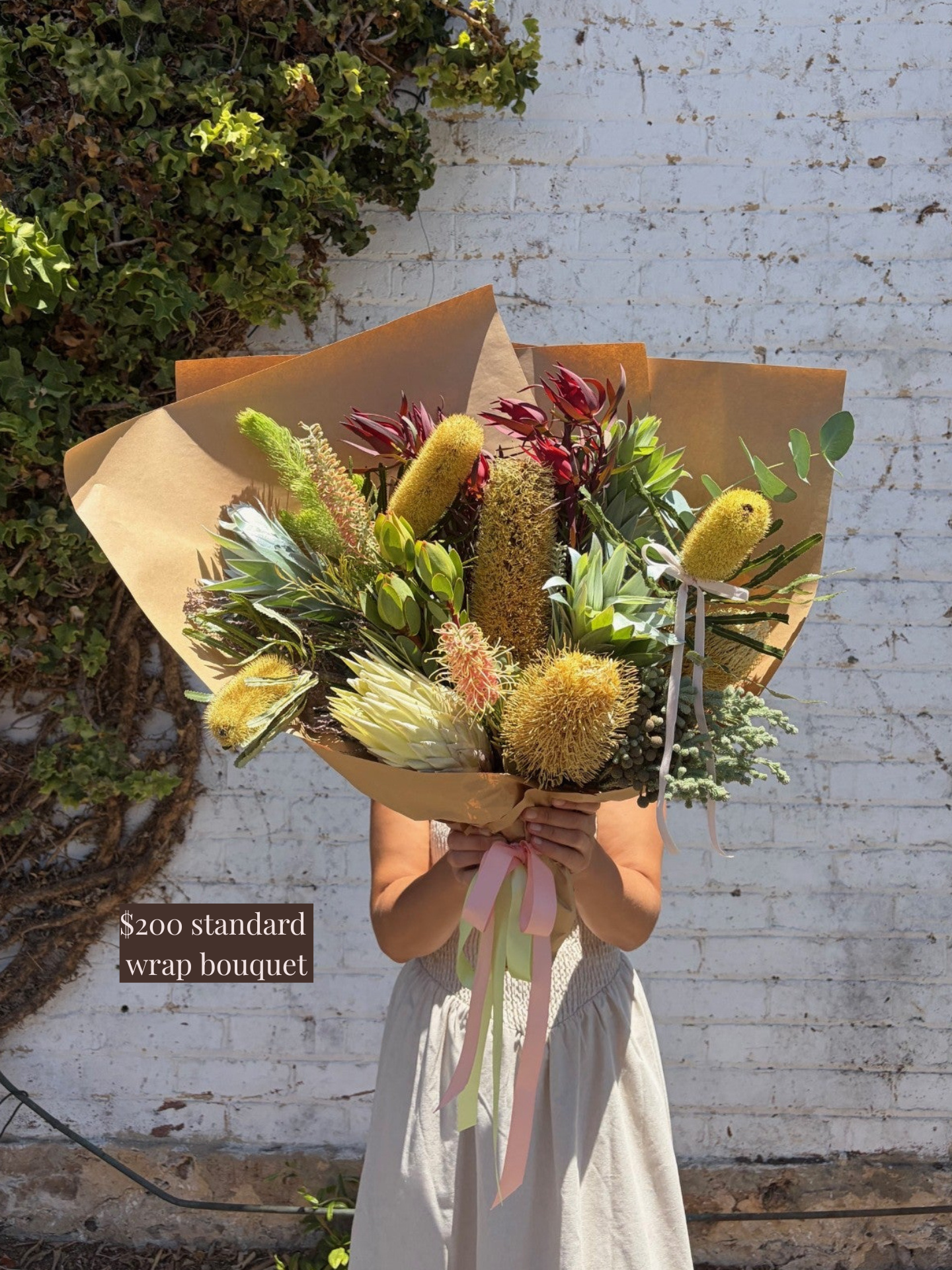 Person holding a bouquet of flowers wrapped in brown paper against a white brick wall.