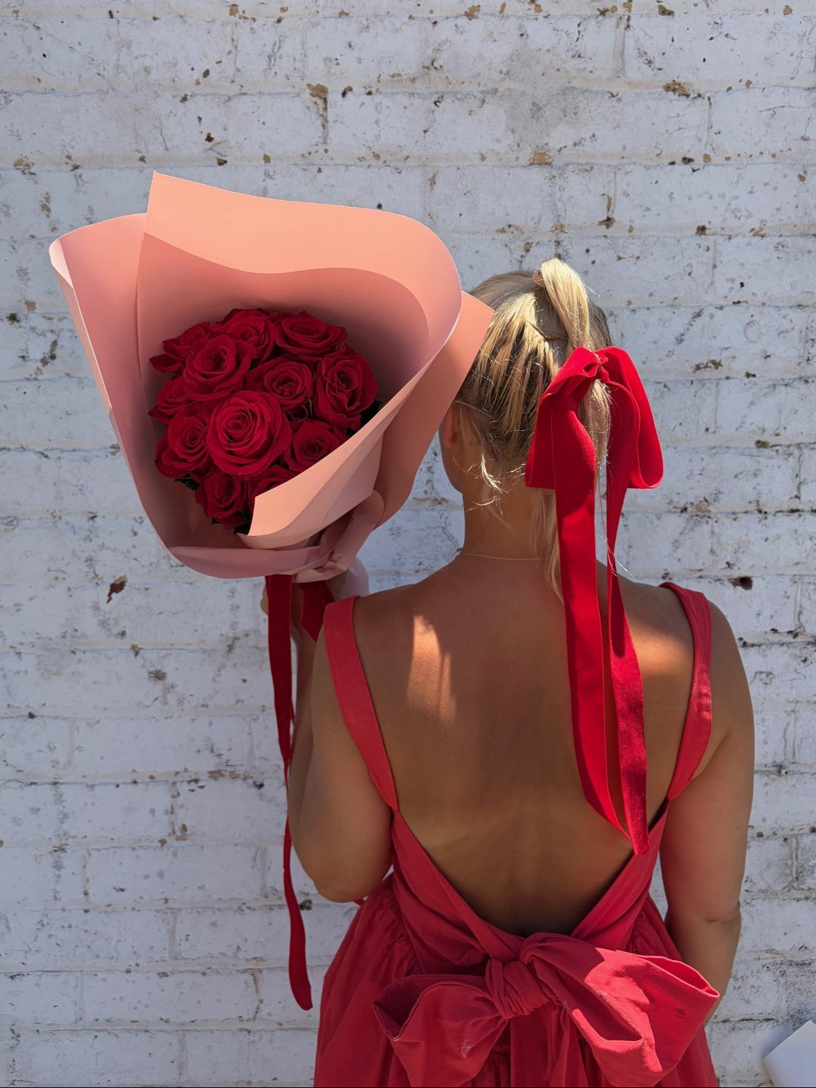 Woman in a red dress holding a large bouquet of red roses against a white brick wall.
