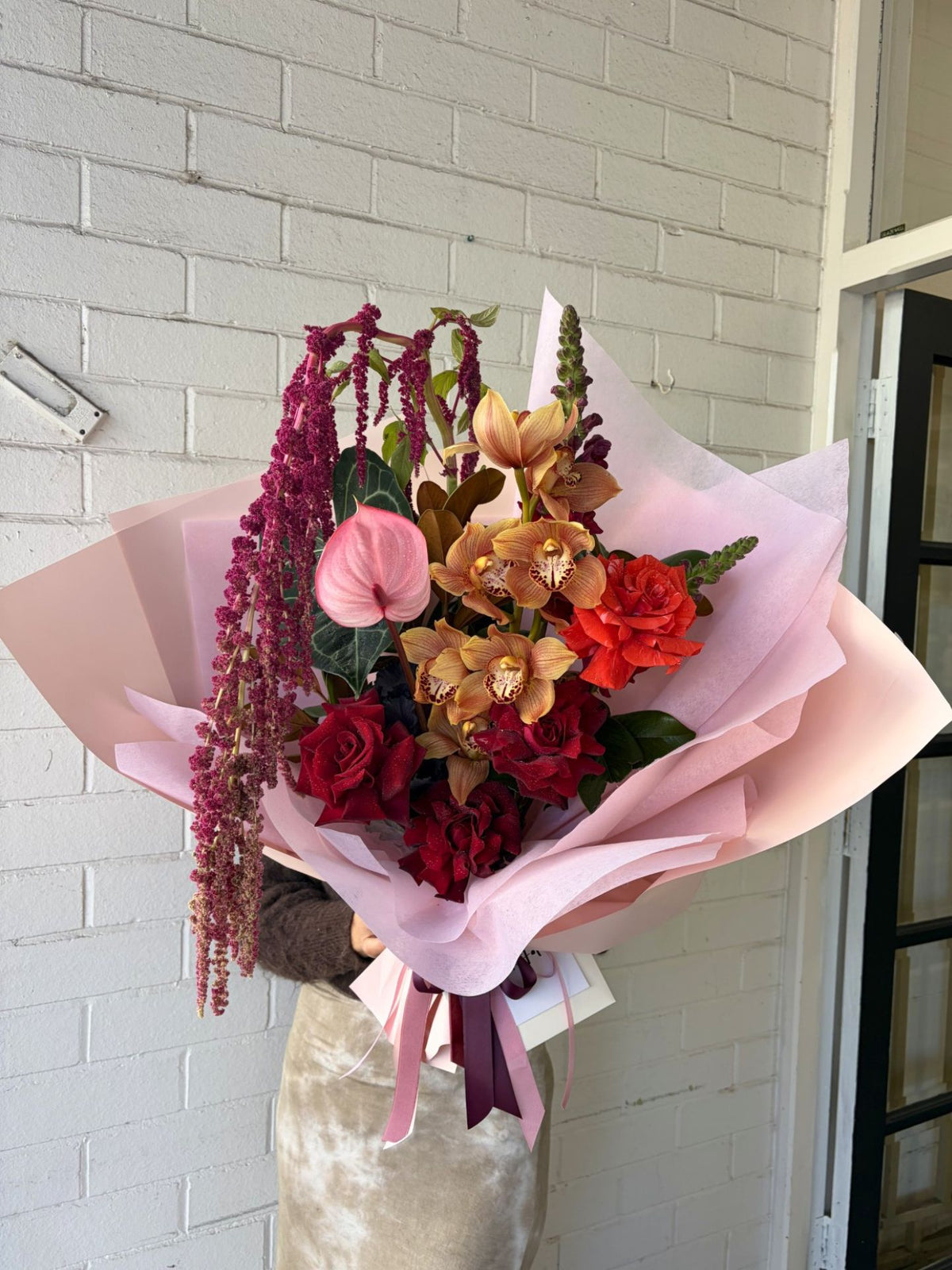 A large, beautifully arranged floral bouquet wrapped in light pink tissue paper, held by a person wearing a beige velvet top. The bouquet features a variety of flowers including red roses, a pink anthurium, orange and yellow orchids, burgundy berries, and green foliage, set against a white brick wall background.