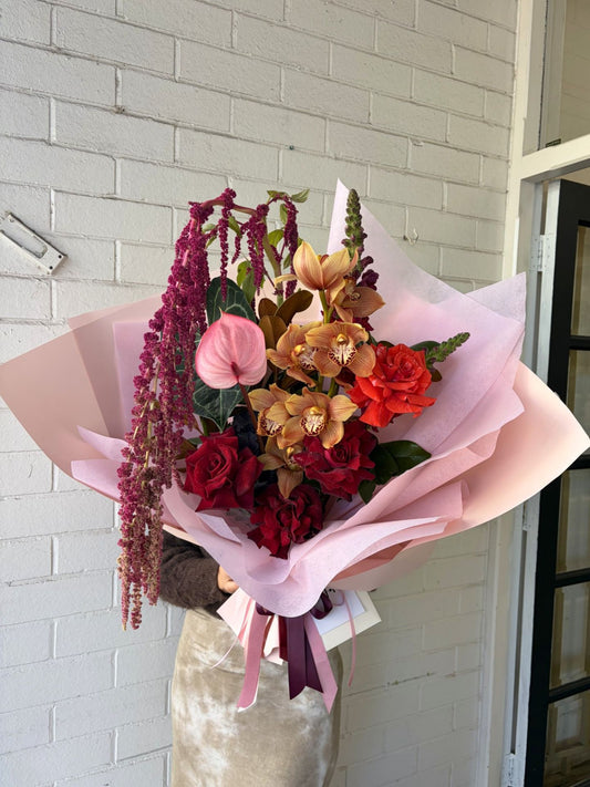 A large, beautifully arranged floral bouquet wrapped in light pink tissue paper, held by a person wearing a beige velvet top. The bouquet features a variety of flowers including red roses, a pink anthurium, orange and yellow orchids, burgundy berries, and green foliage, set against a white brick wall background.