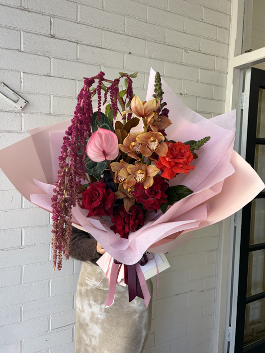 A large, vibrant floral bouquet wrapped in light pink tissue paper, held by a person wearing a beige skirt. The bouquet features a mix of red roses, rust coloured cymbidium orchid, deep burgundy amaranthus, and burgundy snapdragons with green foliage, set against a white painted brick wall.