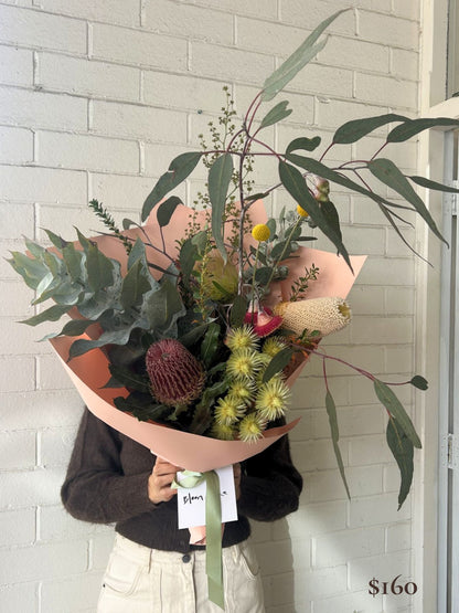 A person in a dark brown sweater and cream pants holds a large, beautifully arranged wildflower bouquet wrapped in peach paper. The bouquet features green eucalyptus leaves, a pinkish-purple protea, yellow spiky flowers, and a cream broomcorn flower, set against a white painted brick wall.