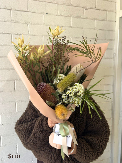A native flower bouquet wrapped in blush pink paper and tied with a sage green ribbon, held by a person wearing a dark brown sherpa jacket against a white painted brick wall. A '$110' price overlay is visible in the bottom-left corner.