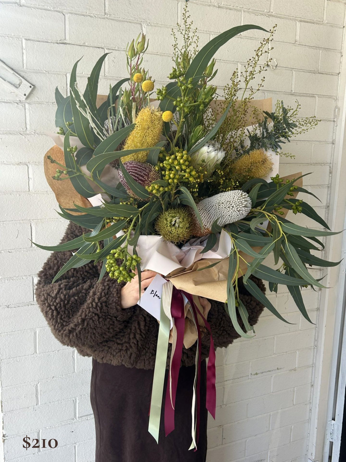 A large, rustic dried and fresh flower bouquet held by a person wearing a brown fuzzy sweater, standing against a white painted brick wall. The bouquet features green eucalyptus leaves, yellow gypsophila, a brown bankia berry, and white protea, wrapped in brown kraft paper with sage green and burgundy ribbons.