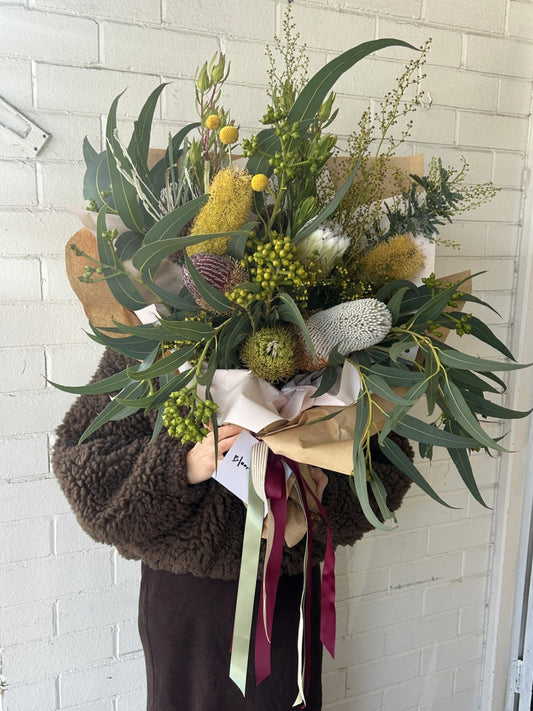 A large, beautifully arranged wildflower bouquet held by a person wearing a brown sherpa jacket. The bouquet features green eucalyptus leaves, yellow and white wildflowers, a green bankweed, and is wrapped in brown kraft paper with cream, sage green, and burgundy ribbons.