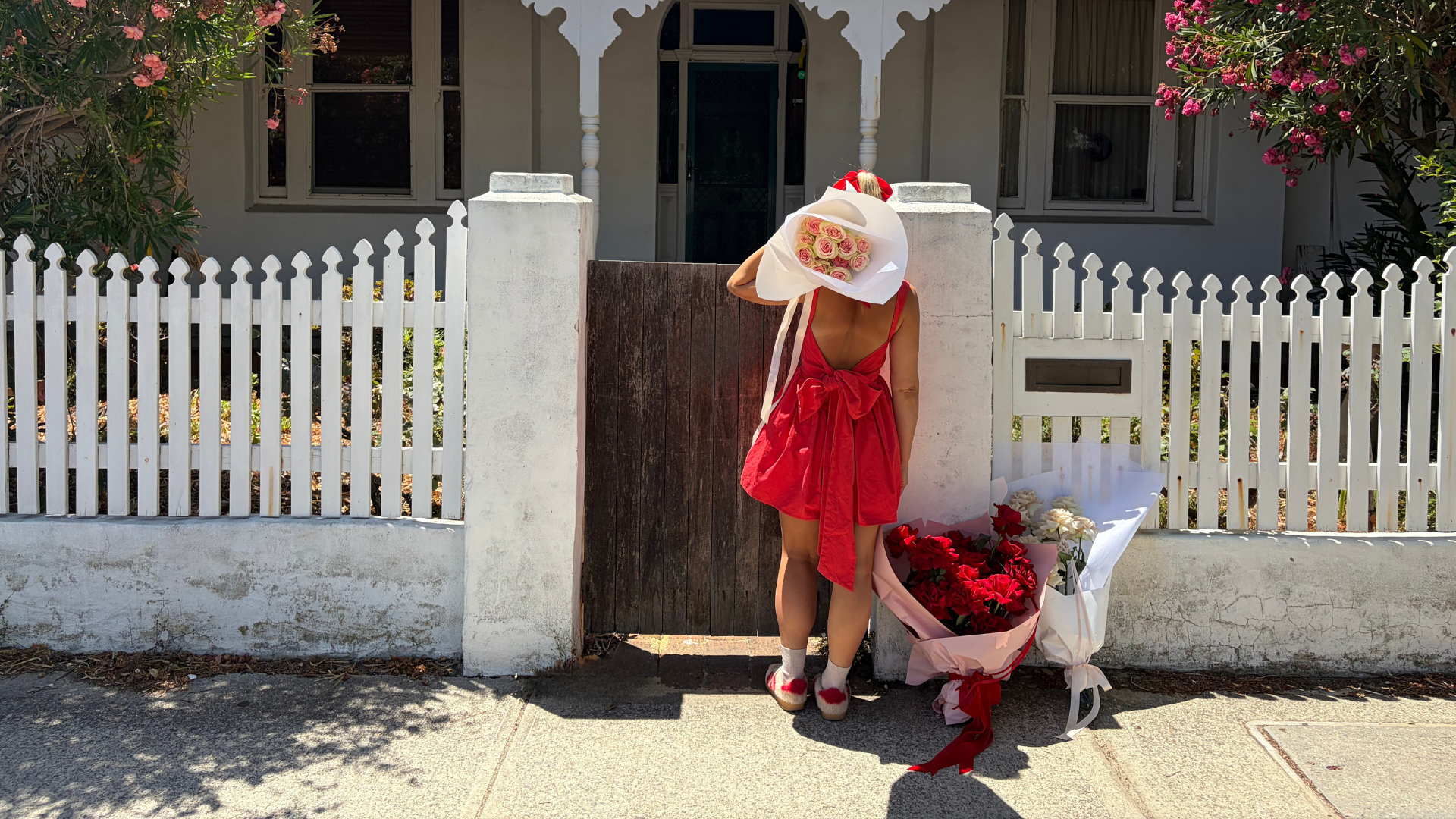 Person in a red dress standing in front of a white picket fence with rose flower bouquets.