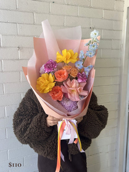 A person in a brown fuzzy jacket holds a colorful bouquet of flowers wrapped in light pink paper, standing against a white brick wall.