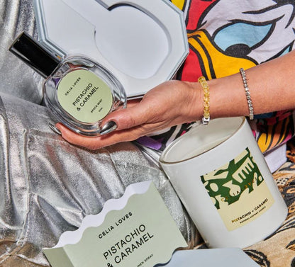 Hand holding a perfume bottle labeled 'Pistachio & Caramel' by Celia Lover, with a candle and packaging in the background.