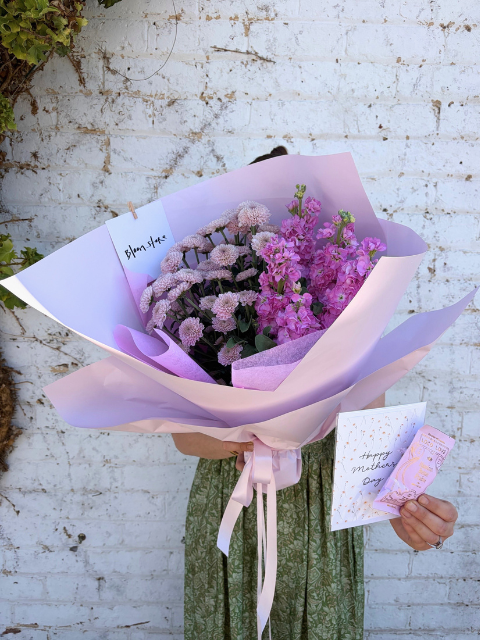 Bouquet of flowers wrapped in purple paper with a card, held against a textured white wall.