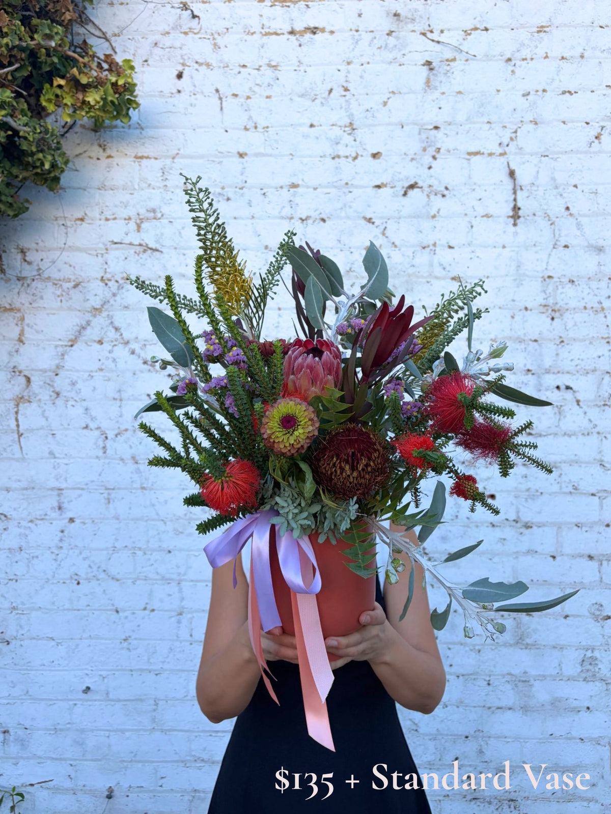 A person holds a large, vibrant floral arrangement in a terracotta-colored pot against a white painted brick wall. The bouquet features a mix of red, pink, yellow, and green flowers with a light purple ribbon.