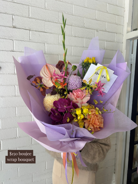Bouquet of flowers wrapped in purple paper with a card, held against a white brick wall.