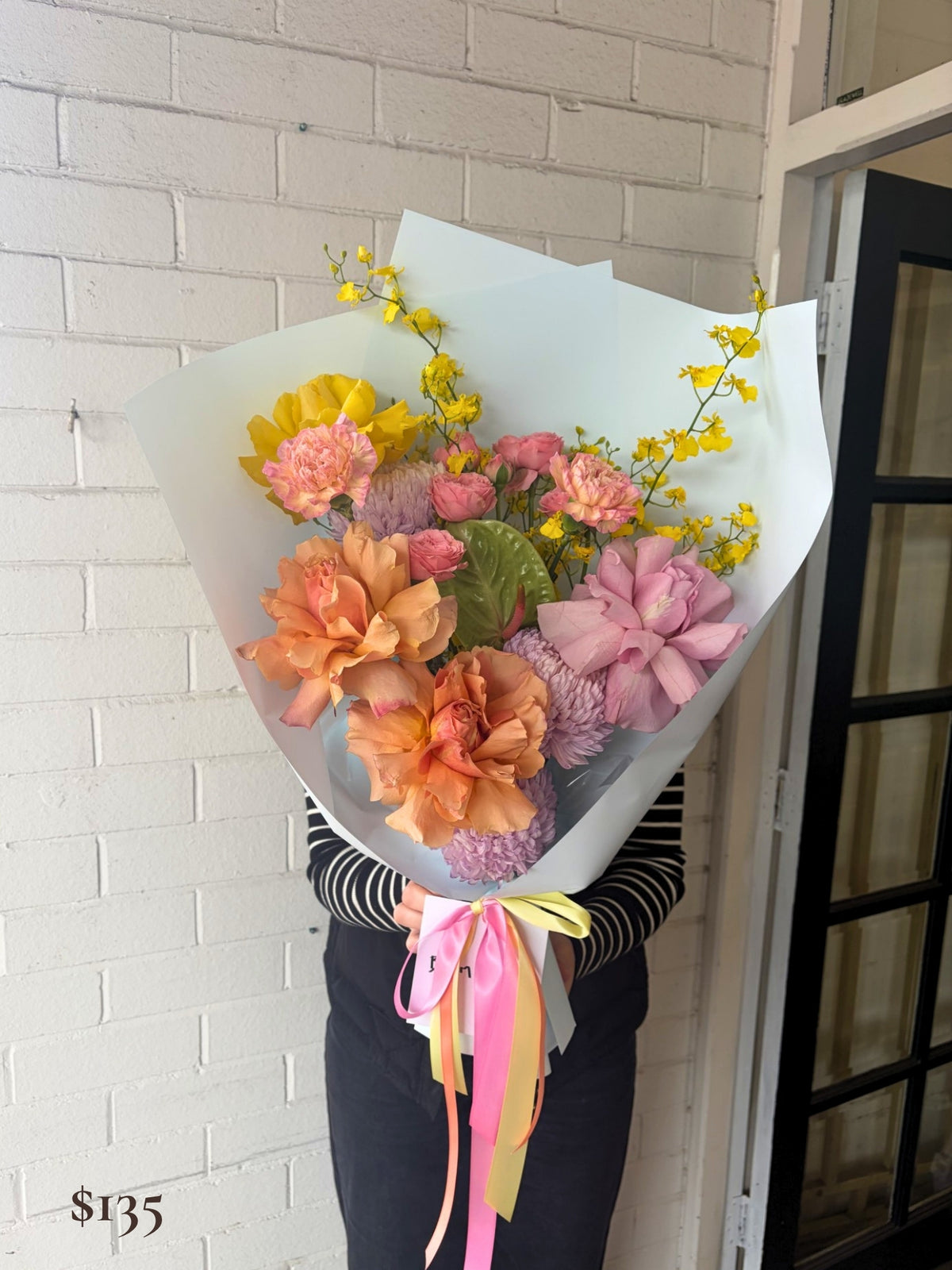 A person holding a large, vibrant flower bouquet wrapped in pale blue paper, featuring pink, peach, yellow, and purple blossoms with colorful ribbons, set against a white brick wall.