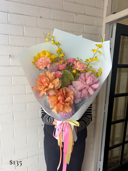 A person holding a large, vibrant flower bouquet wrapped in pale blue paper, featuring pink, peach, yellow, and purple blossoms with colorful ribbons, set against a white brick wall.