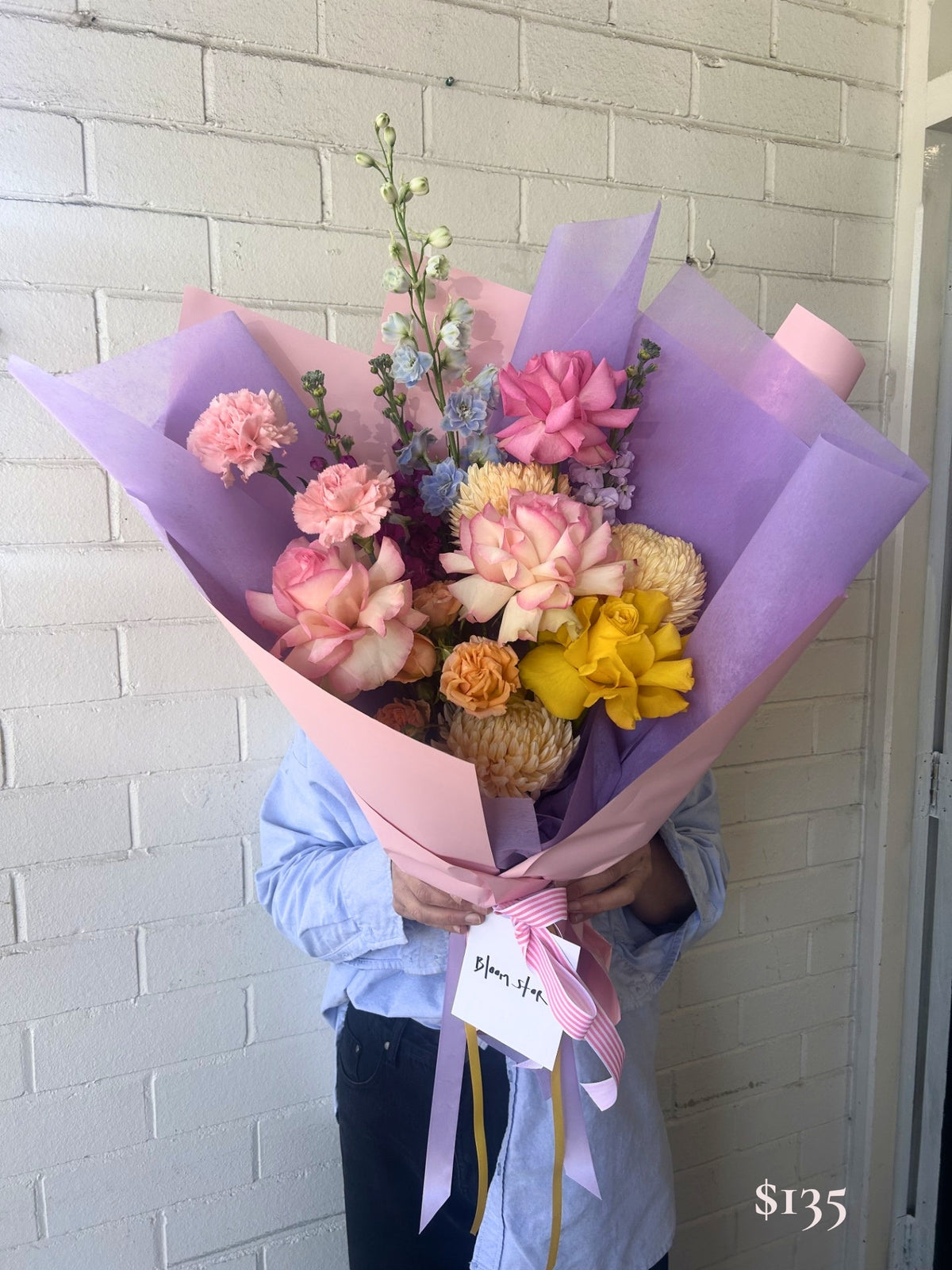 A person in a light blue shirt holds a large bouquet of pink, peach, yellow, and blue flowers wrapped in pink and lavender paper, tied with a pink ribbon and a handwritten gift tag, standing against a white brick wall.