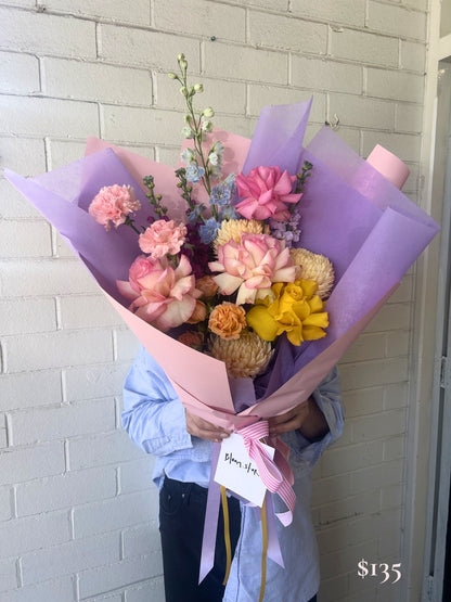 A person in a light blue shirt holds a large bouquet of pink, peach, yellow, and blue flowers wrapped in pink and lavender paper, tied with a pink ribbon and a handwritten gift tag, standing against a white brick wall.