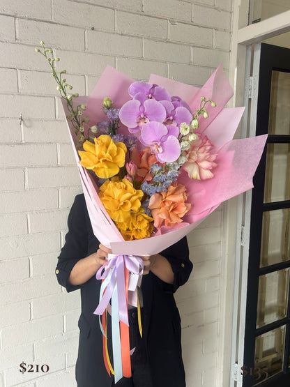 A person in a black coat holds a large, vibrant flower bouquet wrapped in pink paper. The bouquet features purple orchids, yellow carnations, peach roses, and blue filler flowers with pastel ribbons.