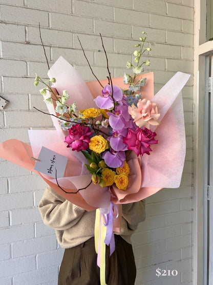 A person holds a large, vibrant floral bouquet wrapped in pink and peach paper with purple and yellow ribbons, standing in front of a white painted brick wall.