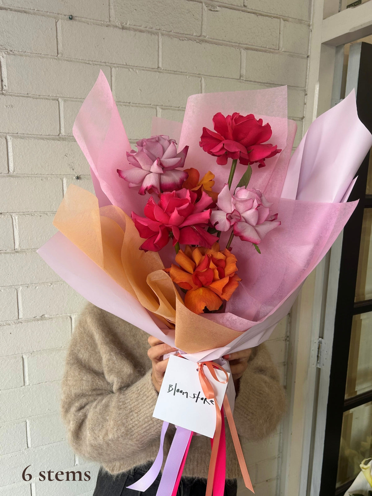 A person in a beige sweater holds a bouquet of artificial flowers wrapped in pink, peach, and white tissue paper with colorful ribbons. The bouquet contains pink, red, and orange flowers, with a white tag reading 'Bloom Staks'.