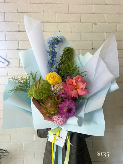 A vibrant mixed flower bouquet wrapped in light blue paper and white tissue, held by a person against a white painted brick wall. The bouquet features a yellow rose, a pink protea, purple flowers, green foliage, and blue delphiniums, with a handwritten price tag in the corner.