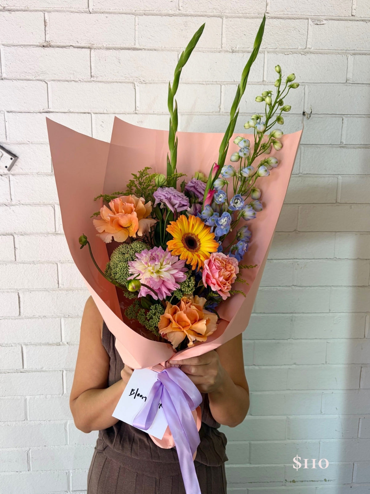 A person holds a large bouquet of colorful flowers wrapped in pink paper with a purple ribbon and a small card, standing against a white painted brick wall.
