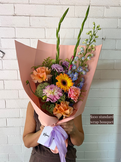 A colourful wrapped flower bouquet held by a person against a white wall.