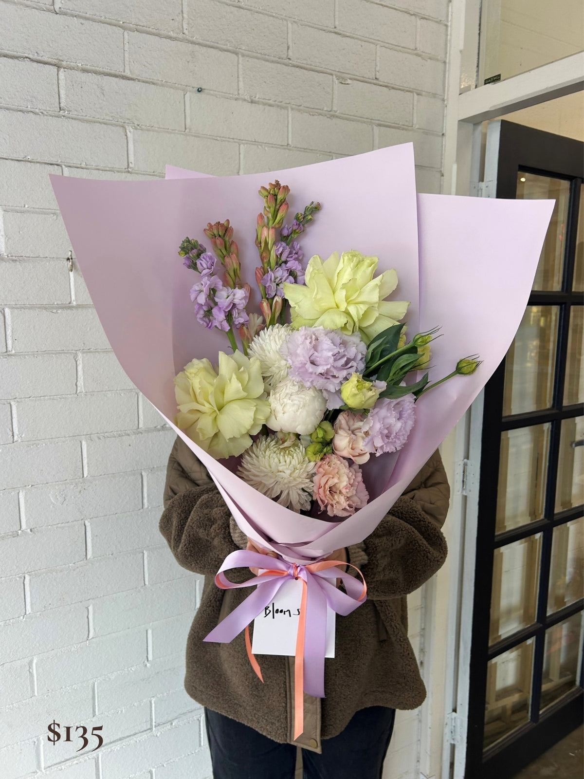 A person in a brown fleece jacket holds a wrapped flower bouquet with pale pink paper wrapping, a pink ribbon, and mixed flowers including pale yellow, lavender, white, and blush pink blooms. The bouquet has a small tag reading 'Blossoms' and is photographed against a white brick wall and a black-framed door.