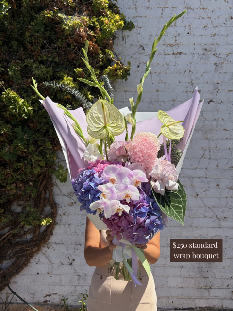 Bouquet of flowers wrapped in purple paper held by a person against a white brick wall.