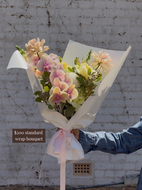 Bouquet of flowers wrapped in white paper with a pink ribbon, held against a textured wall.