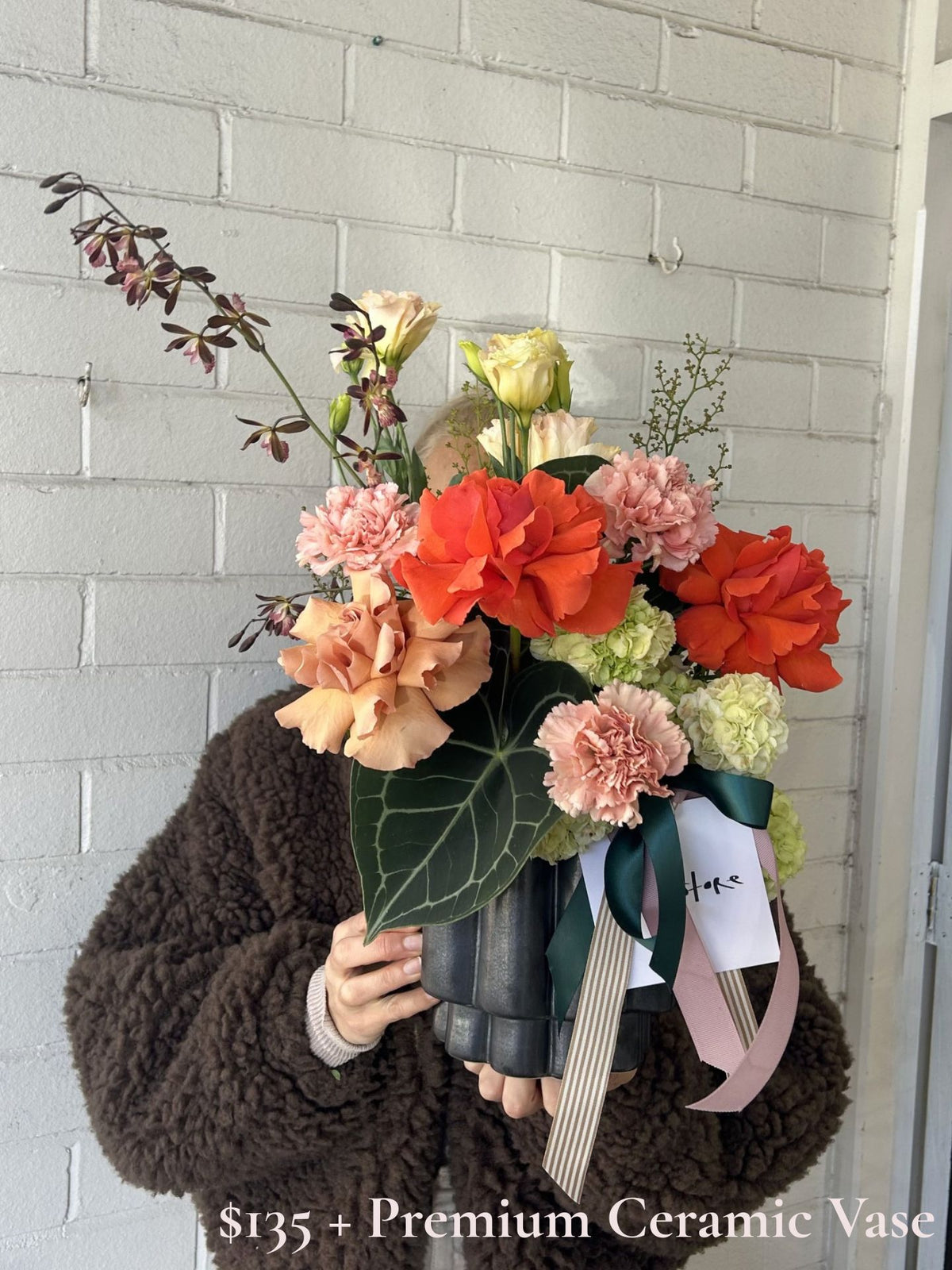 A person in a brown fuzzy jacket holds a large, vibrant flower arrangement in a black ceramic vase against a white painted brick wall. The bouquet features bright orange-red roses, soft pink carnations, peach-colored flowers, and green foliage, tied with dark green and light pink ribbons.