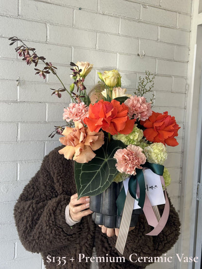 A person in a brown fuzzy jacket holds a large, vibrant flower arrangement in a black ceramic vase against a white painted brick wall. The bouquet features bright orange-red roses, soft pink carnations, peach-colored flowers, and green foliage, tied with dark green and light pink ribbons.