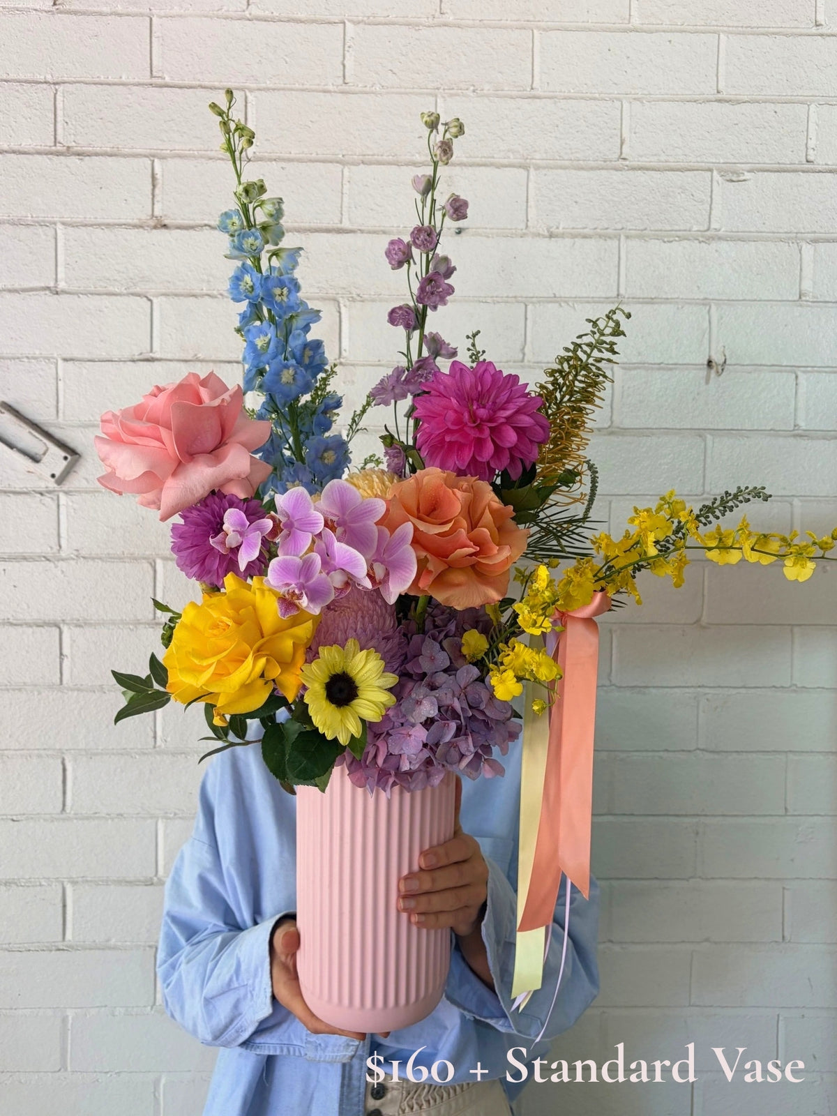 A person in a light blue shirt holds a large, vibrant floral arrangement in a ribbed pink vase against a white brick wall. The bouquet features pink, purple, yellow, and blue flowers with a peach ribbon.