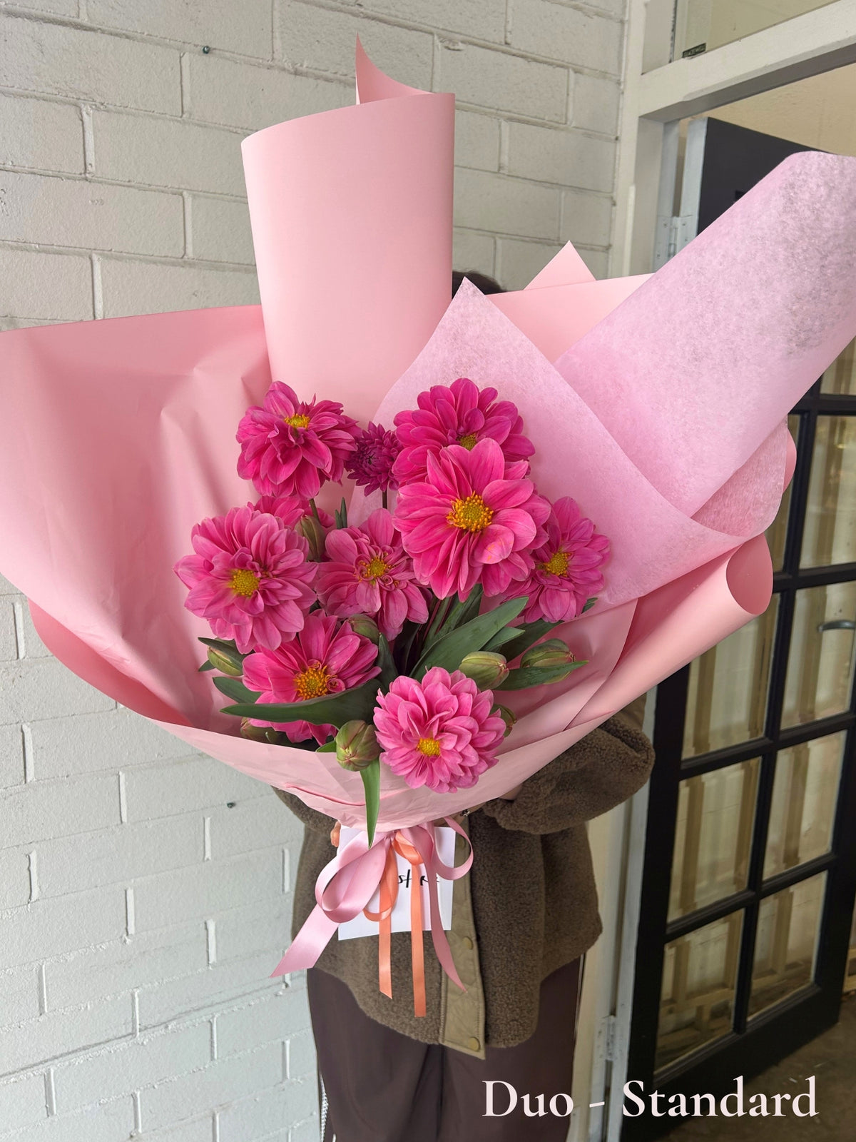 A bouquet of bright pink zinnia flowers wrapped in light pink paper, held by a person wearing a brown jacket, standing in front of a white brick wall and a black-framed glass door.