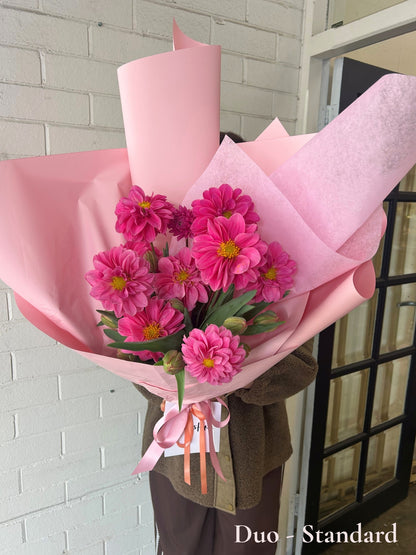 A bouquet of bright pink zinnia flowers wrapped in light pink paper, held by a person wearing a brown jacket, standing in front of a white brick wall and a black-framed glass door.