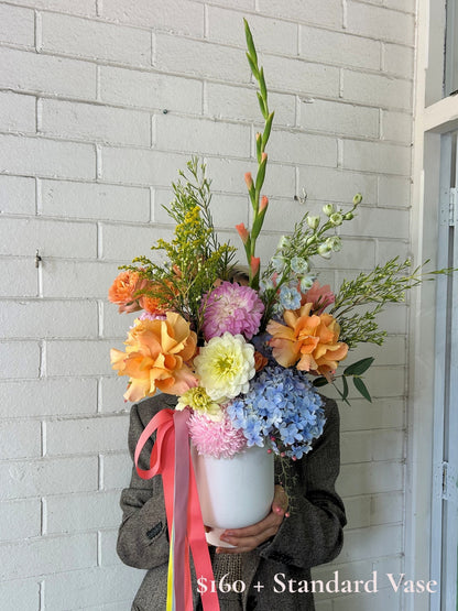 A person in a brown jacket holds a large white ceramic vase filled with a colorful floral arrangement featuring orange, pink, yellow, blue, and green flowers, set against a white painted brick wall.
