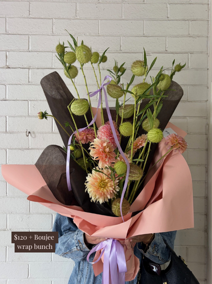 Bouquet of flowers with pink wrap and purple ribbon against a white brick wall.