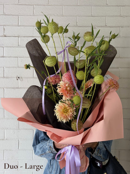 A large flower bouquet wrapped in pink paper and dark brown tissue paper, featuring light green swan plant, pink dahlia, and a purple ribbon, held by a person wearing a denim jacket against a white brick wall.