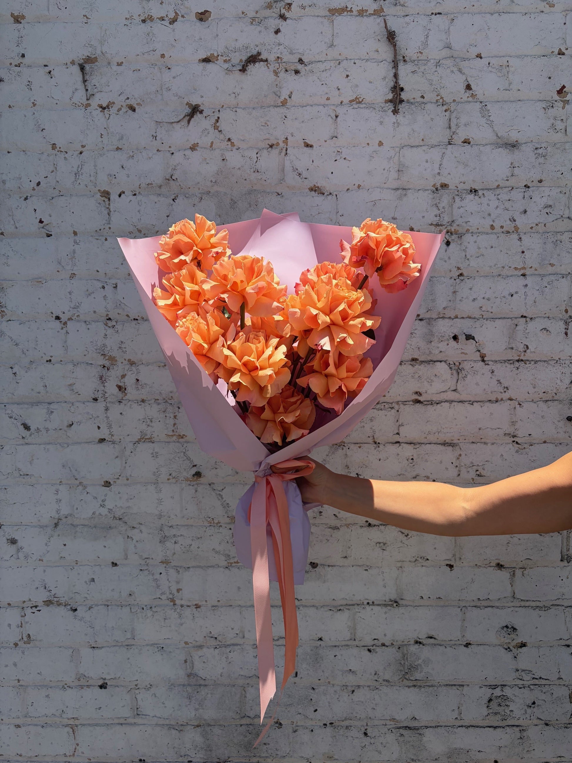 Bouquet of orange flowers wrapped in pink paper held against a white brick wall.