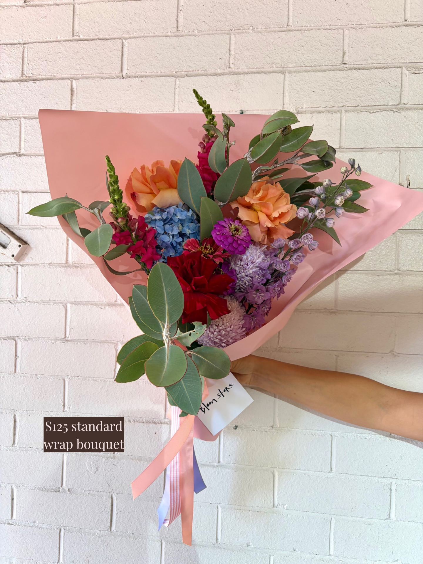 Person holding a bouquet wrapped in pink paper against a white brick wall.