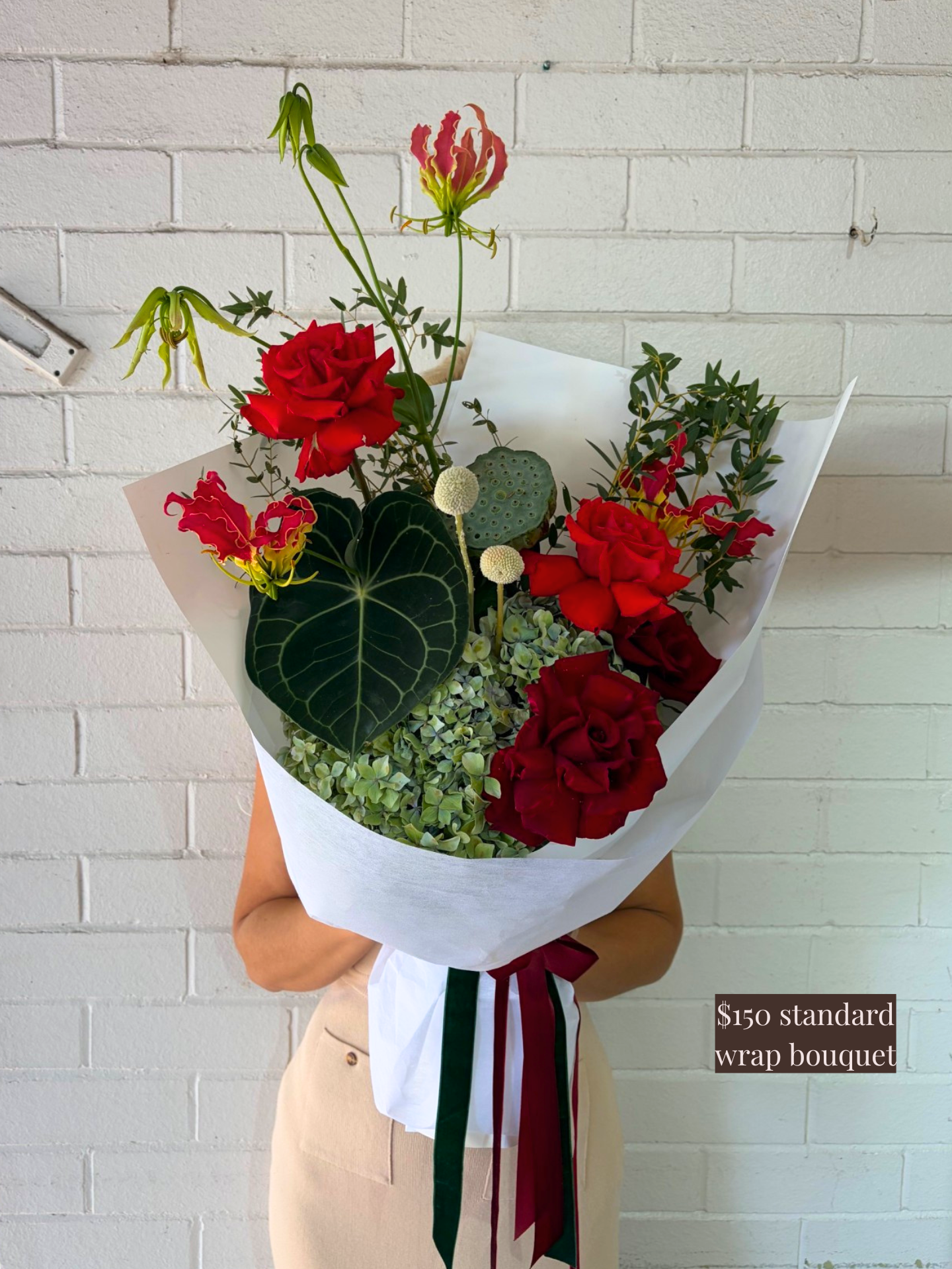 Bouquet of red roses and greenery with a white wrap against a white brick wall.