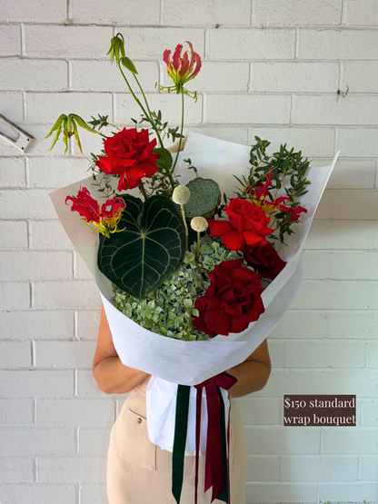 Bouquet of red roses and greenery with a white wrap against a white brick wall.