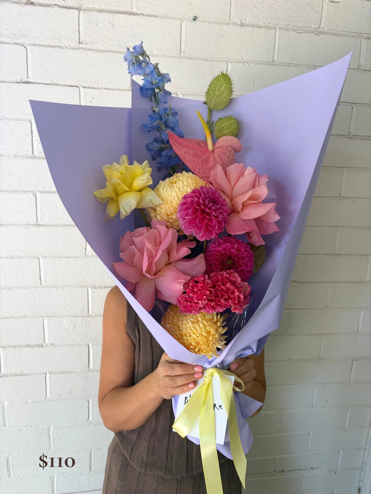A person holding a vibrant mixed flower bouquet wrapped in light purple paper with a pale yellow ribbon, set against a white painted brick wall.
