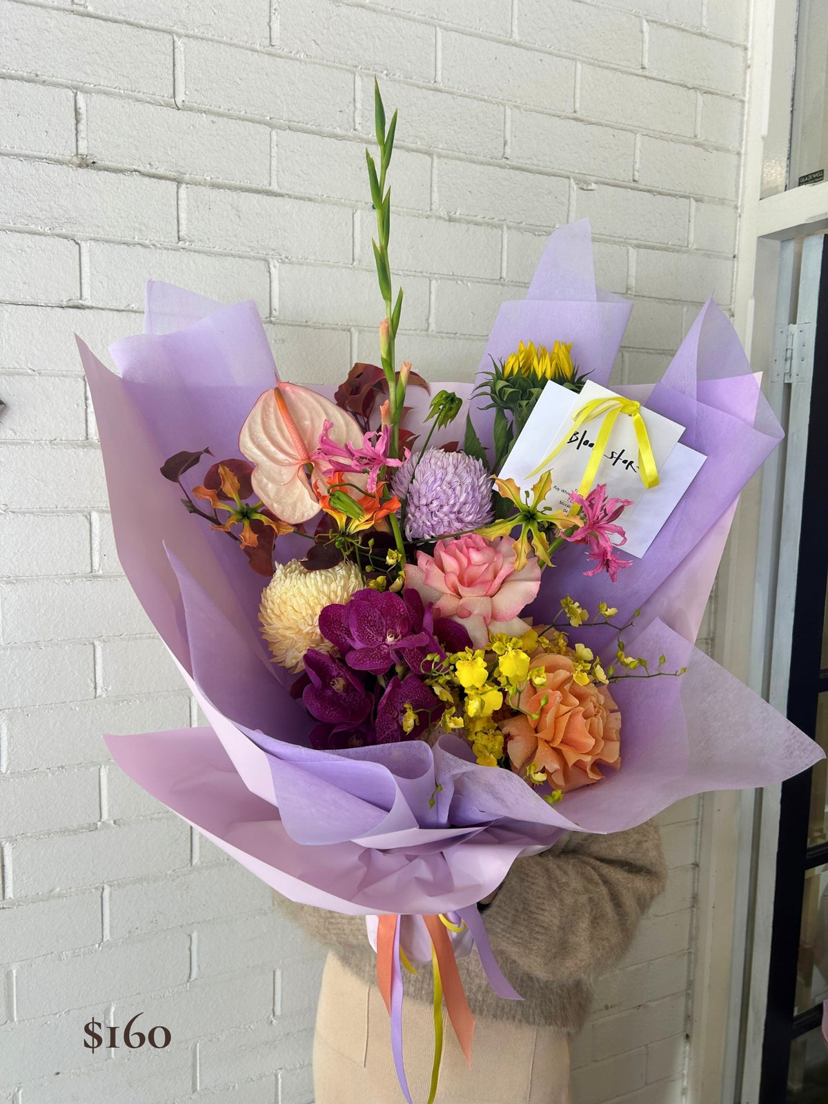 A vibrant mixed flower bouquet wrapped in light purple tissue paper, held by a person wearing a beige sweater. The bouquet features a variety of flowers including pink roses, purple orchids, yellow blossoms, and a white card with a yellow ribbon.