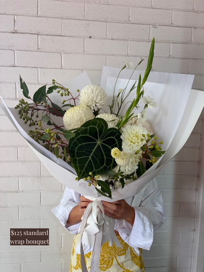 Bouquet of white flowers and greenery wrapped in paper held by a person against a white tiled wall.