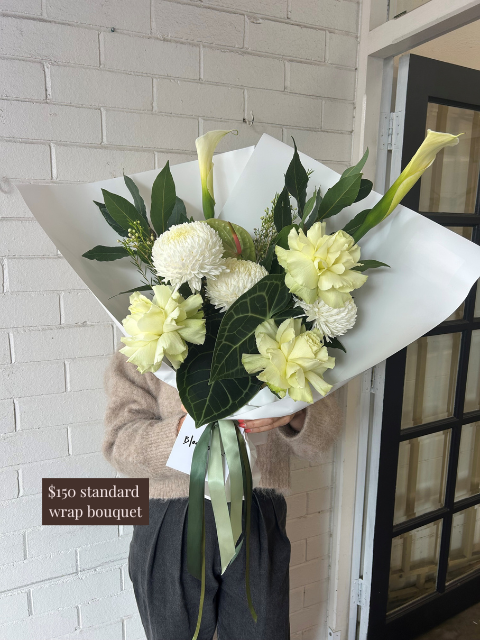 Bouquet of white flowers with green leaves held by a person against a light-colored wall.