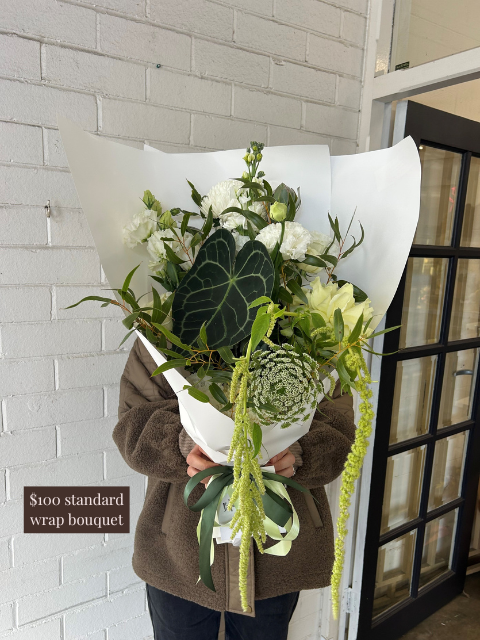 Person holding a bouquet of flowers wrapped in paper against a white brick wall.